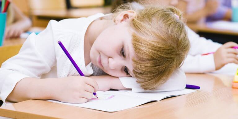 dificuldade de aprendizagem: criança loira escrevendo e com a cabeça deitada na carteira da sala de aula