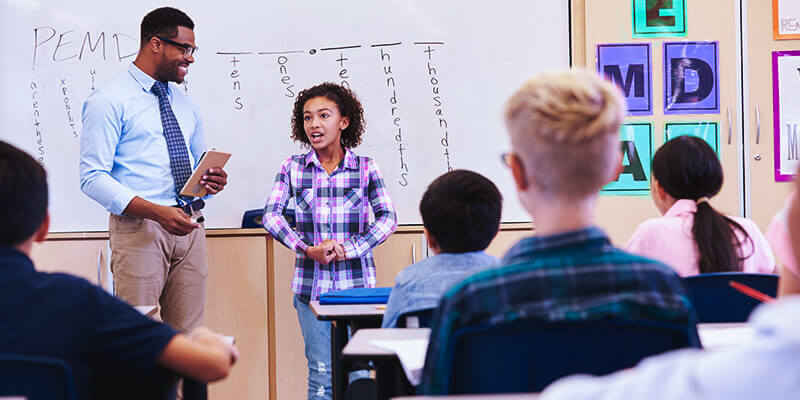Oralidade: criança em sala de aula apresentando na frente dos colegas, ao lado está seu professor