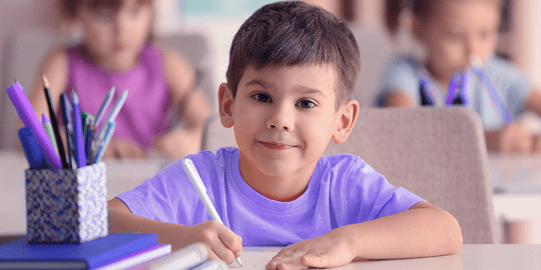 Menino com pequeno sorriso sentado na mesa na escola e escrevendo com um lápis