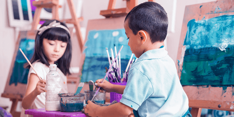 Duas crianças com pincéis escolhendo tinta em uma sala de artes com quadros. Imagem representa um dos tipos de escola fora da linha tradicional