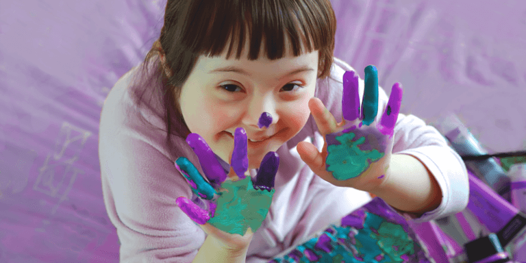 Criança com Síndrome de Down mostrando os dedos sujos de tinta, brincado de pintar e explorando a criatividade infantil