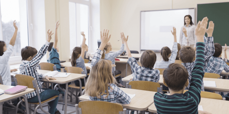 Alunos levantando a mão em sala de aula com professora na frente do quadro