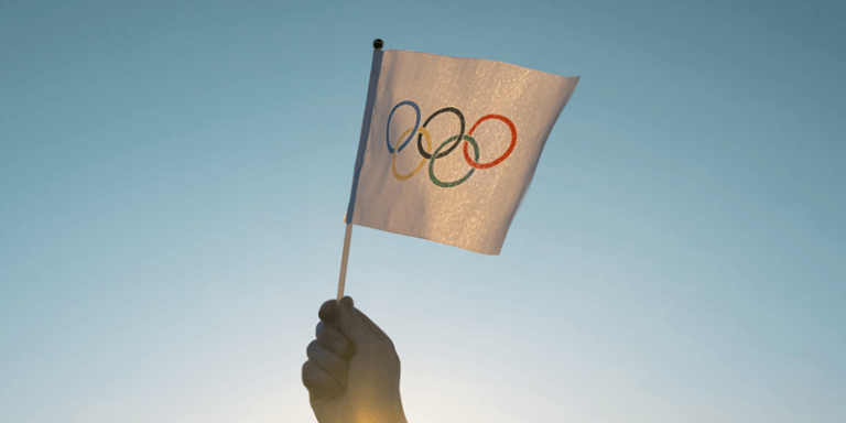 Imagem de uma mão segurando uma bandeira pequena das Olimpíadas com o céu ao fundo