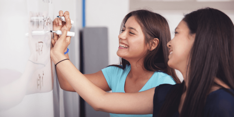 Imagem de duas alunas adolescentes sorrindo e resolvendo exercício na lousa branca